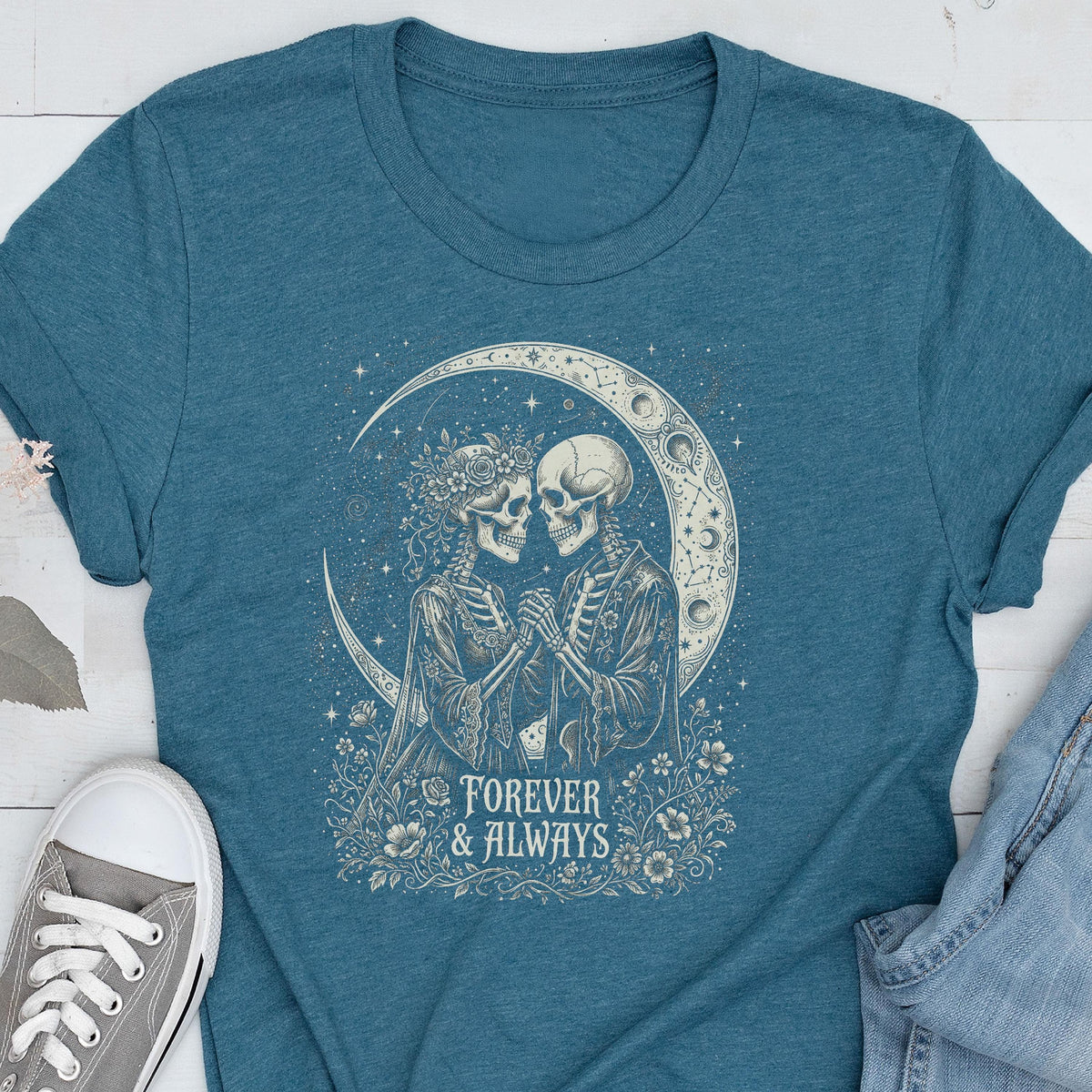 May include: A teal short-sleeve t-shirt featuring a graphic print of two skeletons holding hands in front of a crescent moon. The design includes floral accents and the text "FOREVER & ALWAYS" in a decorative font centered below the skeletons.