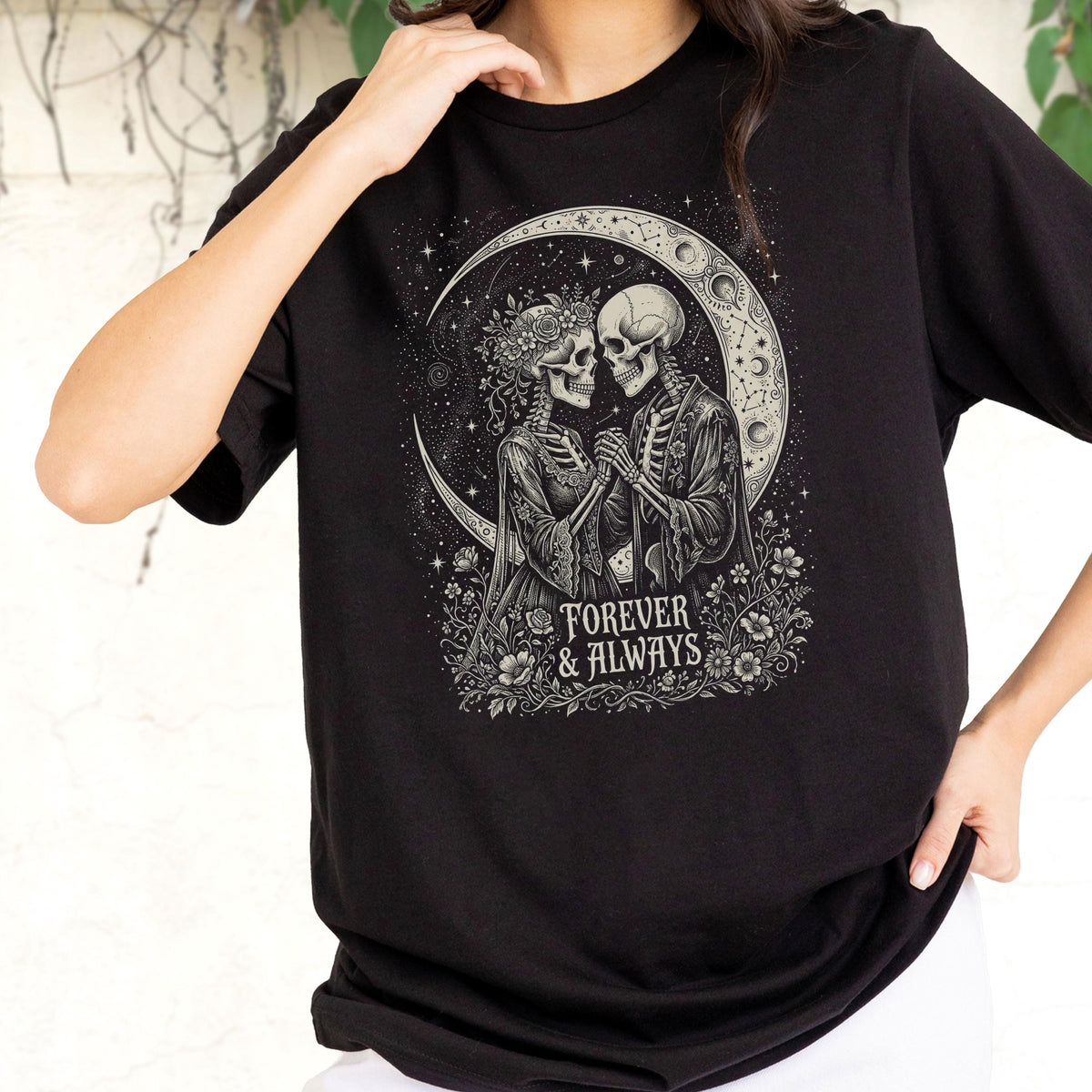 May include: A black t-shirt featuring a graphic print of two skeletons facing each other, holding hands, and surrounded by flowers and a crescent moon. The text "FOREVER & ALWAYS" is printed in a white serif font below the skeletons.