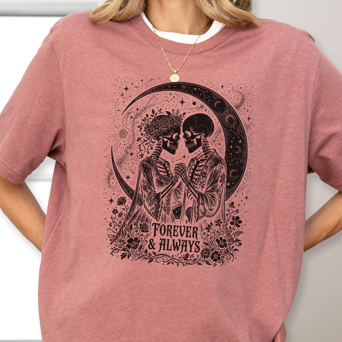May include: A dusty rose t-shirt featuring a black graphic print of two skeletons holding hands under a crescent moon. The design includes floral accents and the text "FOREVER & ALWAYS" in a serif font, surrounded by stars and celestial patterns.
