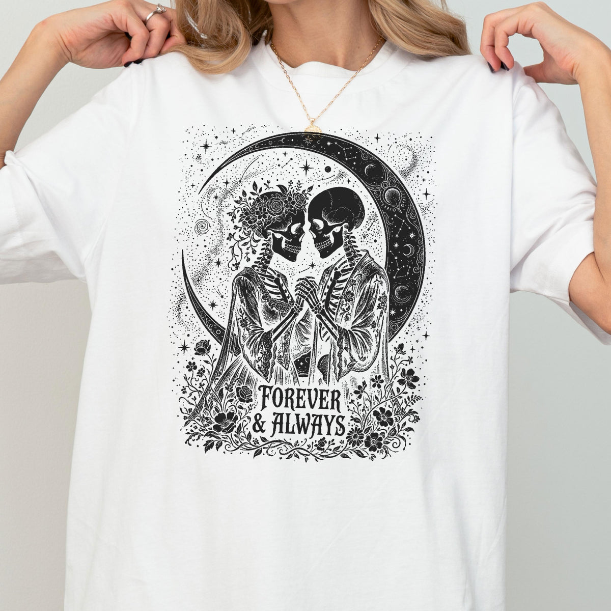 May include: A white t-shirt featuring a black graphic print of two skeletons facing each other, adorned with floral crowns and robes. The design includes a large crescent moon, stars, and the text Forever & Always surrounded by flowers.