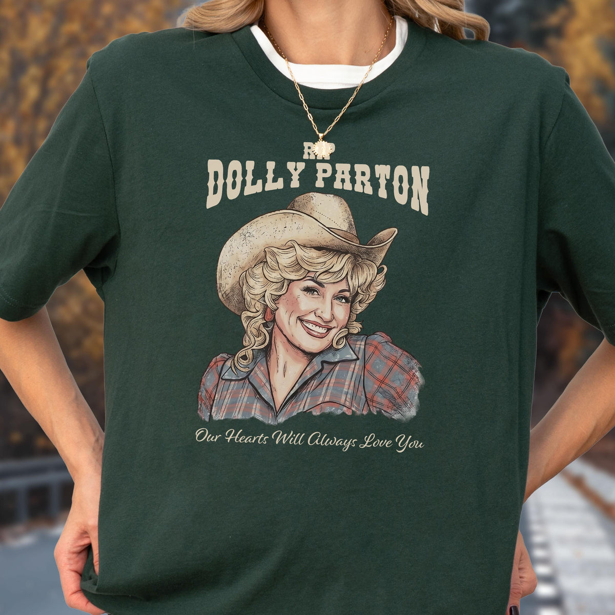 May include: A dark green short-sleeved t-shirt featuring a graphic illustration of Dolly Parton wearing a cowboy hat and a plaid shirt. The text reads "RIP DOLLY PARTON" above the portrait and "Our Hearts Will Always Love You" below it.