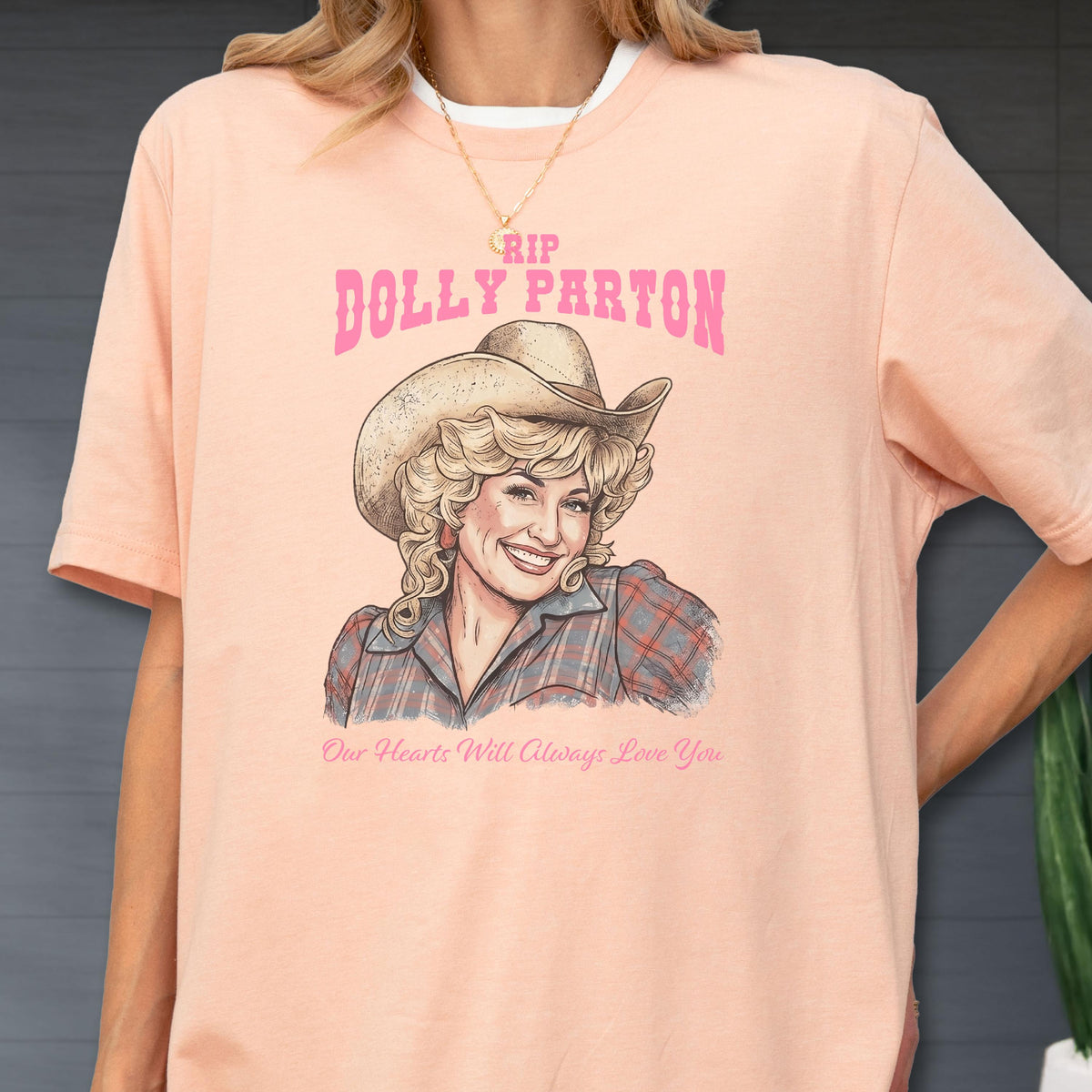 May include: A peach-colored t-shirt featuring a graphic illustration of Dolly Parton wearing a cowboy hat and a plaid shirt. The text above reads RIP DOLLY PARTON in pink, and the text below reads Our Hearts Will Always Love You in pink.