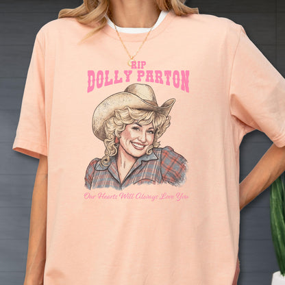 May include: A peach-colored t-shirt featuring a graphic illustration of Dolly Parton wearing a cowboy hat and a plaid shirt. The text above reads RIP DOLLY PARTON in pink, and the text below reads Our Hearts Will Always Love You in pink.