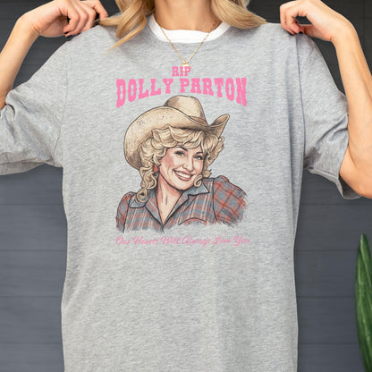 May include: A heather gray t-shirt featuring a graphic print of Dolly Parton wearing a cowboy hat and a plaid shirt. Above the portrait, the text reads RIP DOLLY PARTON in pink, and below, the text reads Our Hearts Will Always Love You.