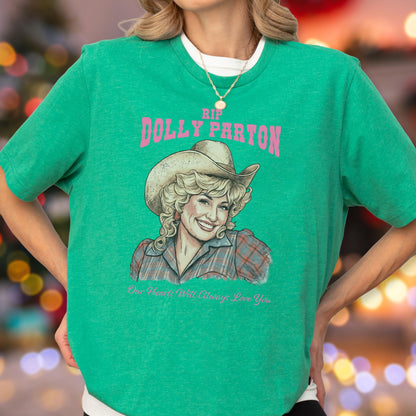 May include: A bright green short-sleeved t-shirt featuring a graphic illustration of Dolly Parton wearing a cowboy hat and a plaid shirt. The text reads RIP DOLLY PARTON in pink letters above the portrait and Our Hearts Will Always Love You below.