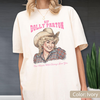 May include: An ivory short-sleeve t-shirt featuring a graphic illustration of Dolly Parton wearing a cowboy hat and a plaid shirt. The text reads RIP DOLLY PARTON in pink letters above the illustration and Our Hearts Will Always Love You below.