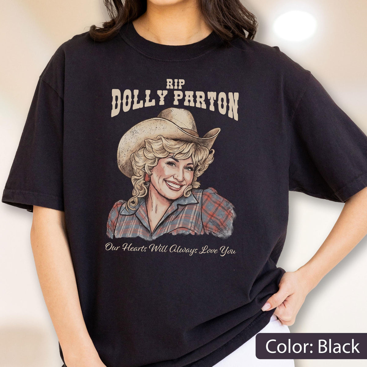 May include: A black t-shirt featuring a graphic illustration of Dolly Parton wearing a cowboy hat and a plaid shirt. The text above reads RIP DOLLY PARTON and the text below reads Our Hearts Will Always Love You in a cursive script font.