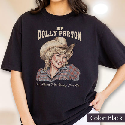 May include: A black t-shirt featuring a graphic illustration of Dolly Parton wearing a cowboy hat and a plaid shirt. The text above reads RIP DOLLY PARTON and the text below reads Our Hearts Will Always Love You in a cursive script font.