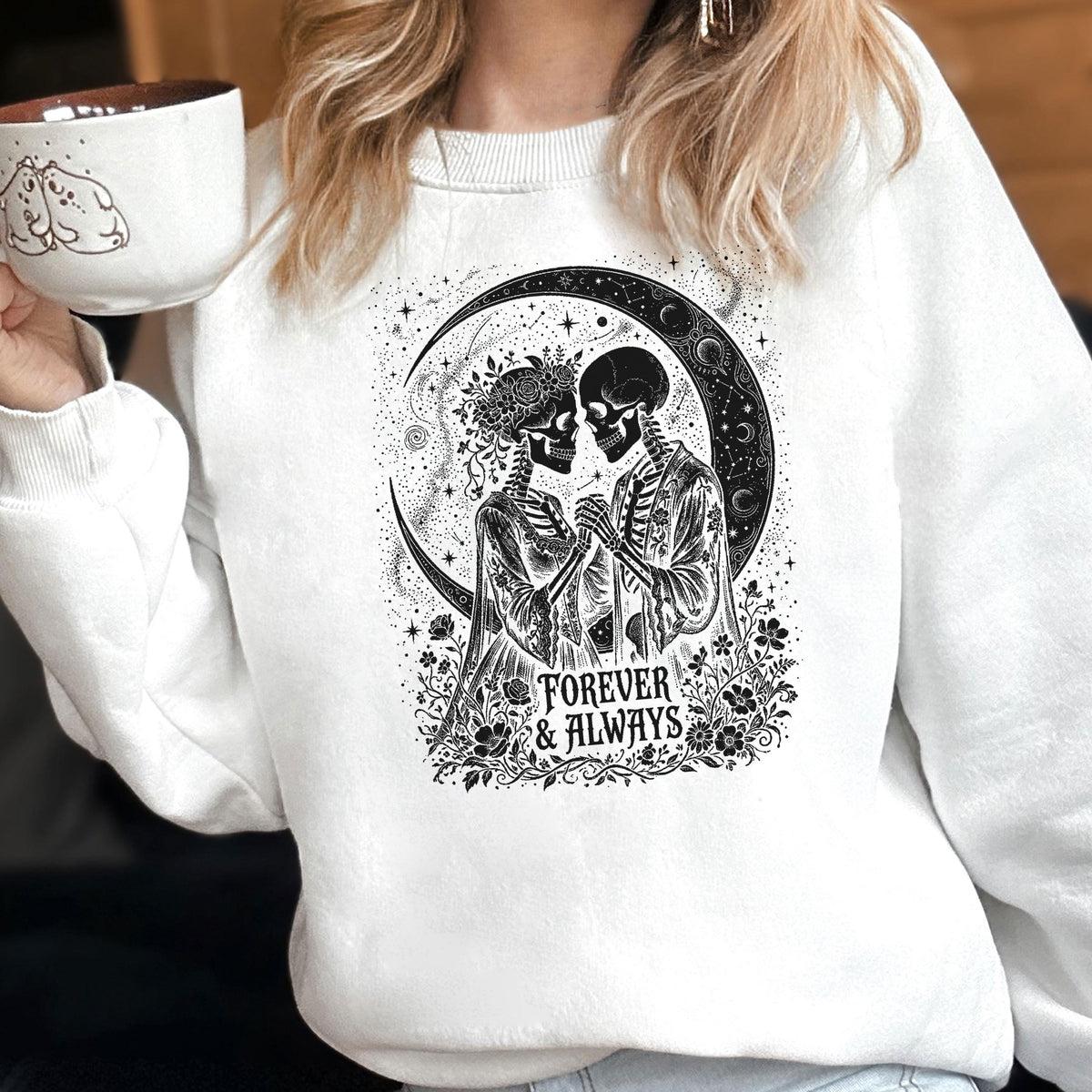 May include: A white crewneck sweatshirt featuring a black graphic print of two skeletons holding hands in front of a crescent moon. The design includes floral accents and the text Forever & Always. A white mug with a small black line drawing is visible.