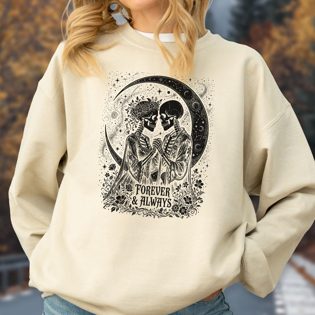 May include: A beige crewneck sweatshirt featuring a black graphic print of two skeletons holding hands under a crescent moon. The design includes floral accents and the text "FOREVER & ALWAYS" centered at the bottom of the illustration.