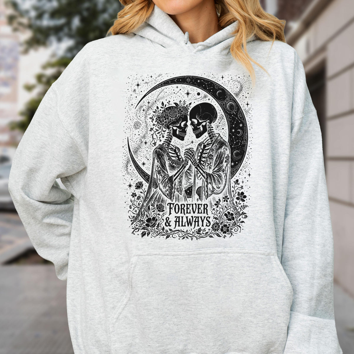 May include: A light gray hooded sweatshirt featuring a black graphic print of two skeletons holding hands under a crescent moon. The design includes floral elements and the text "FOREVER & ALWAYS" centered at the bottom of the illustration.