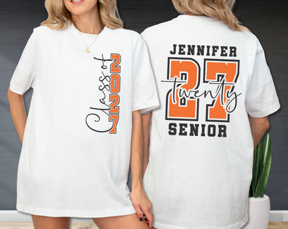 May include: A white t-shirt featuring a graphic design on the front and back. The front displays the text "Class of 2027" in black and orange. The back features the name "JENNIFER" above the number "27" in orange, with "twenty" and "SENIOR" text.