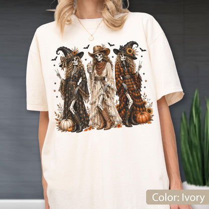 May include: An ivory t-shirt featuring a graphic print of three skeletons dressed in western-style clothing and hats. The skeletons are surrounded by pumpkins, sunflowers, and bats. The design includes intricate details on their dresses and boots.