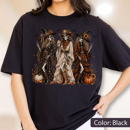 May include: A black t-shirt featuring a graphic print of three skeletons dressed in western-style clothing and hats. The skeletons are surrounded by pumpkins, sunflowers, and small bats, with one skeleton holding a cup and others making peace signs.