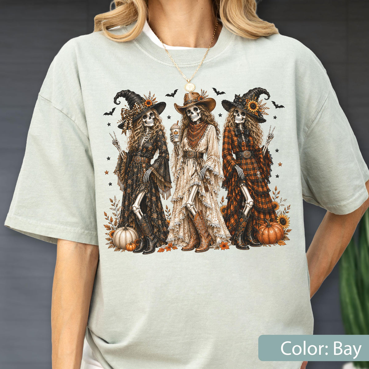 May include: A light sage green t-shirt featuring a graphic print of three skeletons dressed in western-style witch attire, including hats, long dresses, and boots, surrounded by pumpkins, sunflowers, and bats, with a gold necklace worn above.