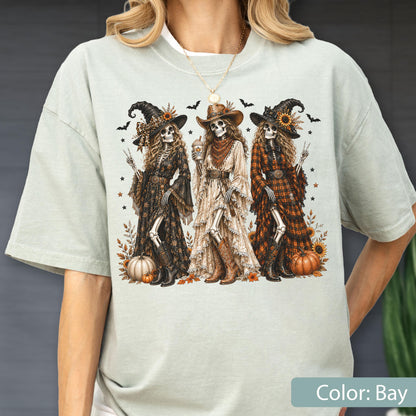 May include: A light sage green t-shirt featuring a graphic print of three skeletons dressed in western-style witch attire, including hats, long dresses, and boots, surrounded by pumpkins, sunflowers, and bats, with a gold necklace worn above.