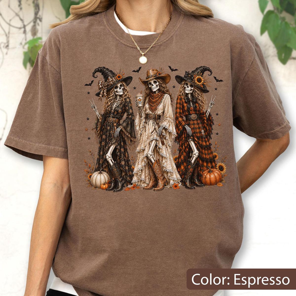 May include: An espresso-colored t-shirt featuring a graphic print of three skeletons dressed in western-style clothing and hats. The skeletons are surrounded by pumpkins, sunflowers, and bats. The shirt has a crew neckline and short sleeves.