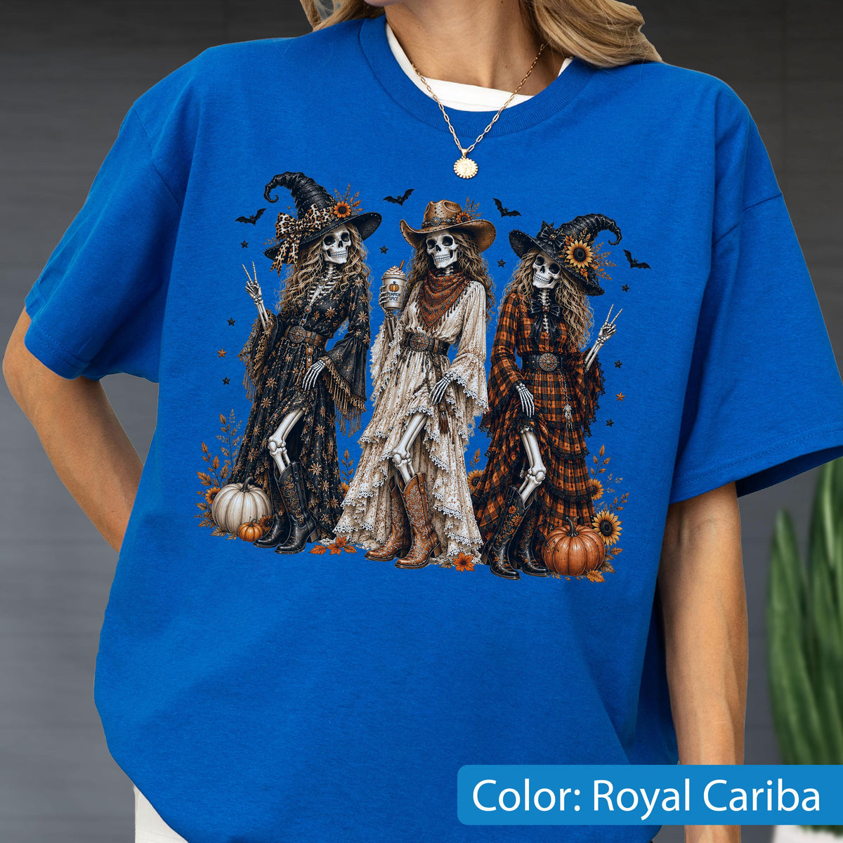 May include: A royal blue t-shirt featuring a graphic print of three skeletons dressed in western-style clothing, including hats, dresses, and boots. The skeletons are surrounded by pumpkins, sunflowers, and bats. The text Color: Royal Cariba is visible.