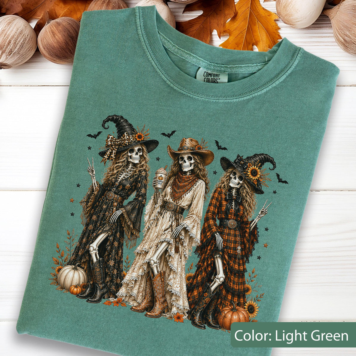 May include: A light green t-shirt featuring a graphic of three skeletons dressed in western-style clothing and hats. The skeletons are surrounded by pumpkins, sunflowers, and bats. The shirt has a Comfort Colors brand label at the inner neckline.