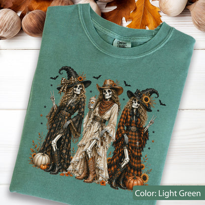 May include: A light green t-shirt featuring a graphic of three skeletons dressed in western-style clothing and hats. The skeletons are surrounded by pumpkins, sunflowers, and bats. The shirt has a Comfort Colors brand label at the inner neckline.