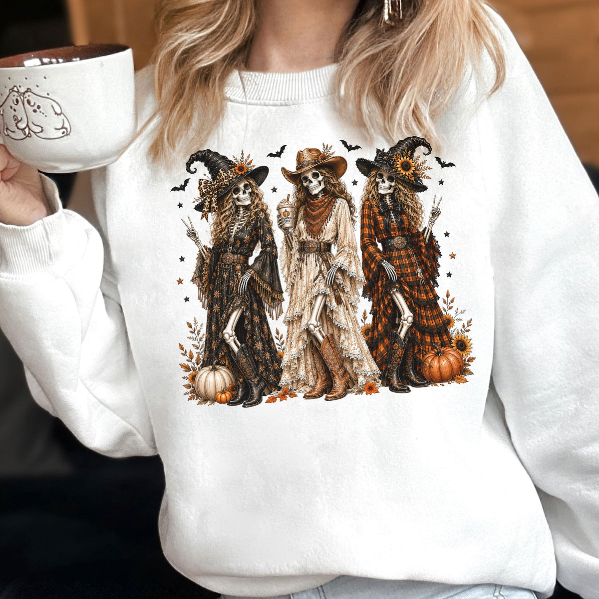 May include: A white crewneck sweatshirt featuring a graphic print of three skeletons dressed in western-style clothing, including hats, long dresses, and boots. The skeletons are surrounded by pumpkins, sunflowers, bats, and autumn leaves.
