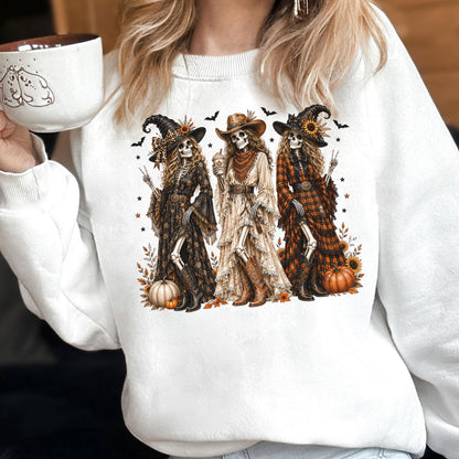 May include: A white crewneck sweatshirt featuring a graphic print of three skeletons dressed in western-style clothing, including hats, long dresses, and boots. The skeletons are surrounded by pumpkins, sunflowers, bats, and autumn leaves.