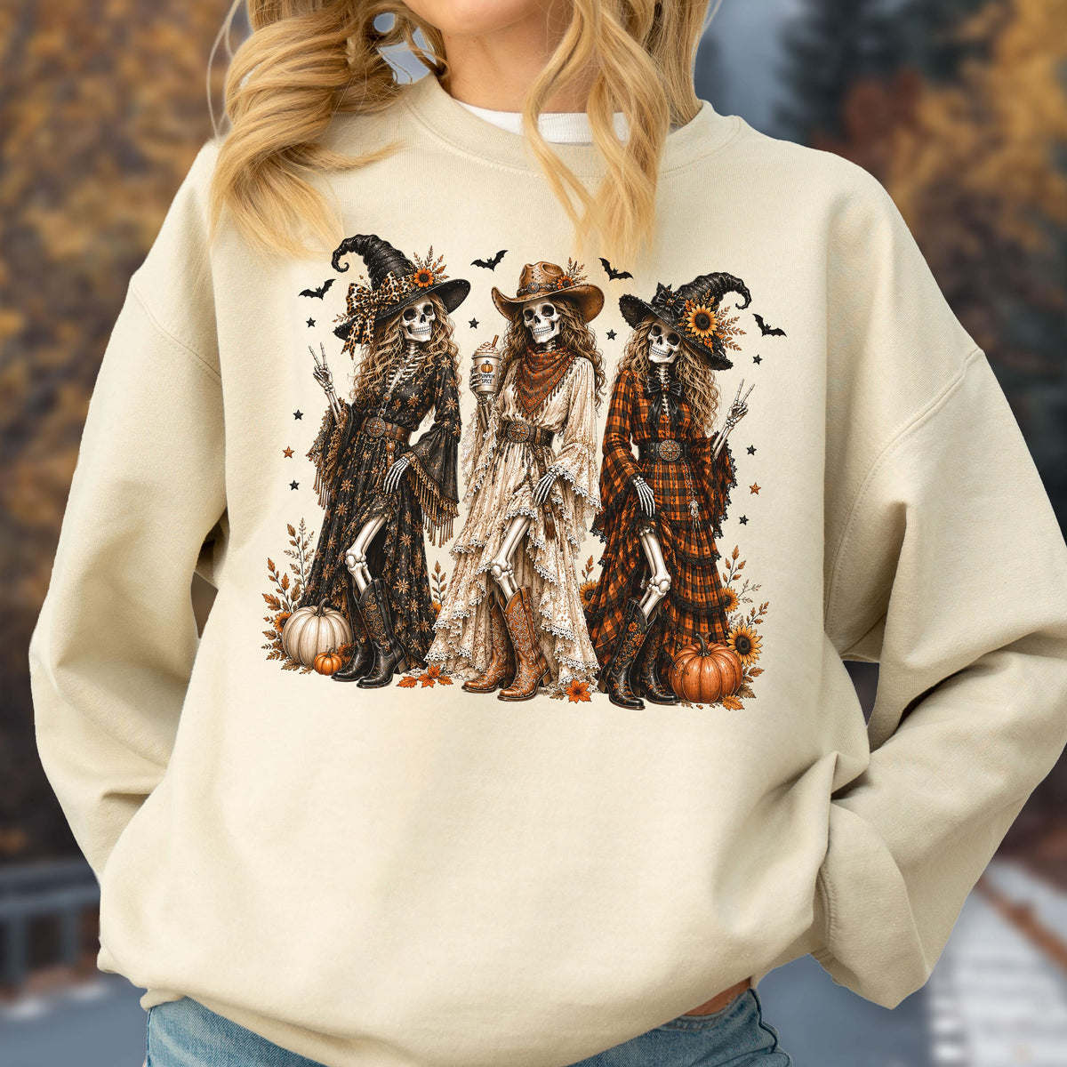 May include: A beige crewneck sweatshirt featuring a graphic print of three skeletons dressed in western-style clothing, including witch hats, cowboy hats, and boots. The skeletons are surrounded by pumpkins, sunflowers, and small flying bats.