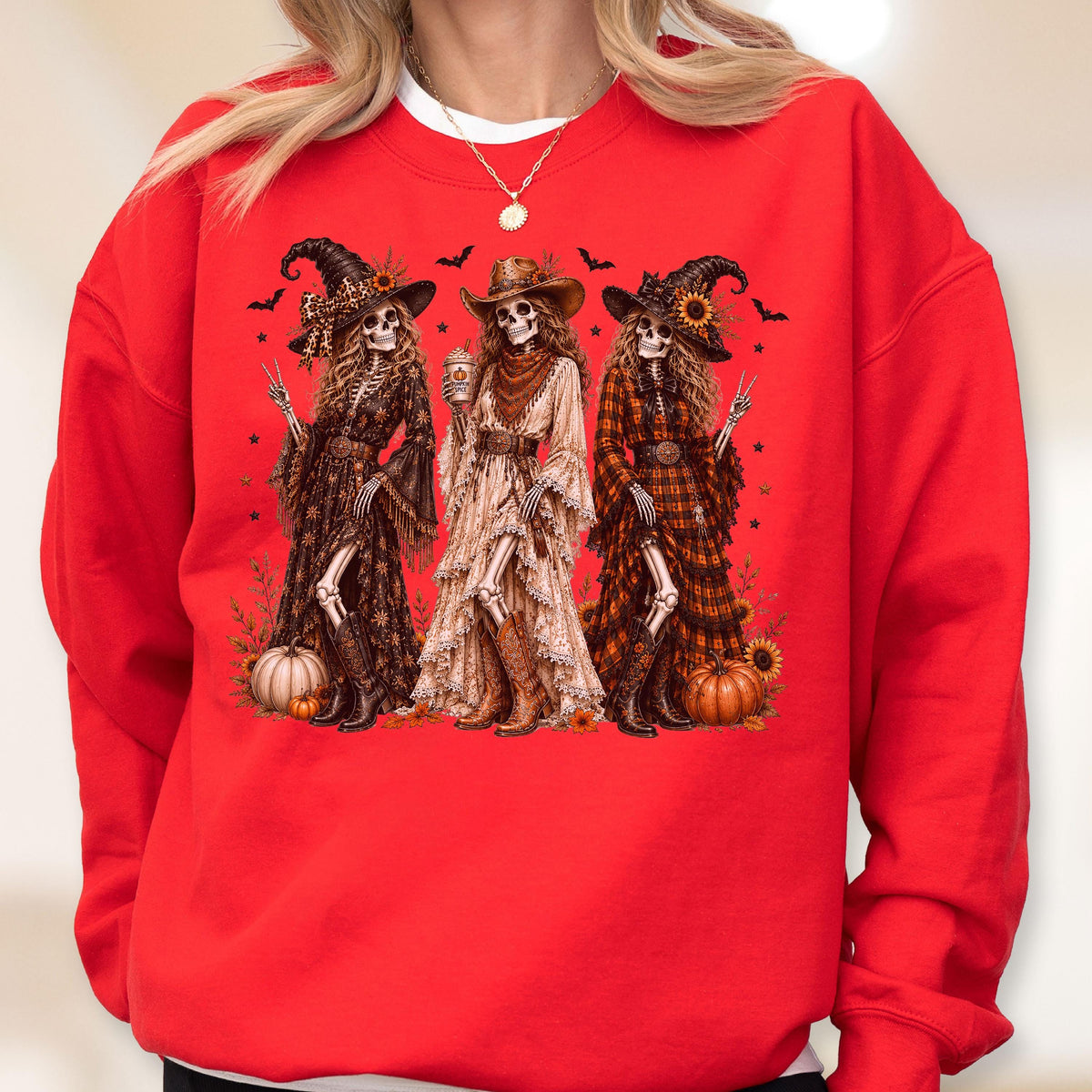 May include: A bright red crewneck sweatshirt featuring a graphic print of three skeleton figures dressed in western-style hats, long dresses, and boots. The skeletons are surrounded by pumpkins, sunflowers, and small bats on a solid red background.