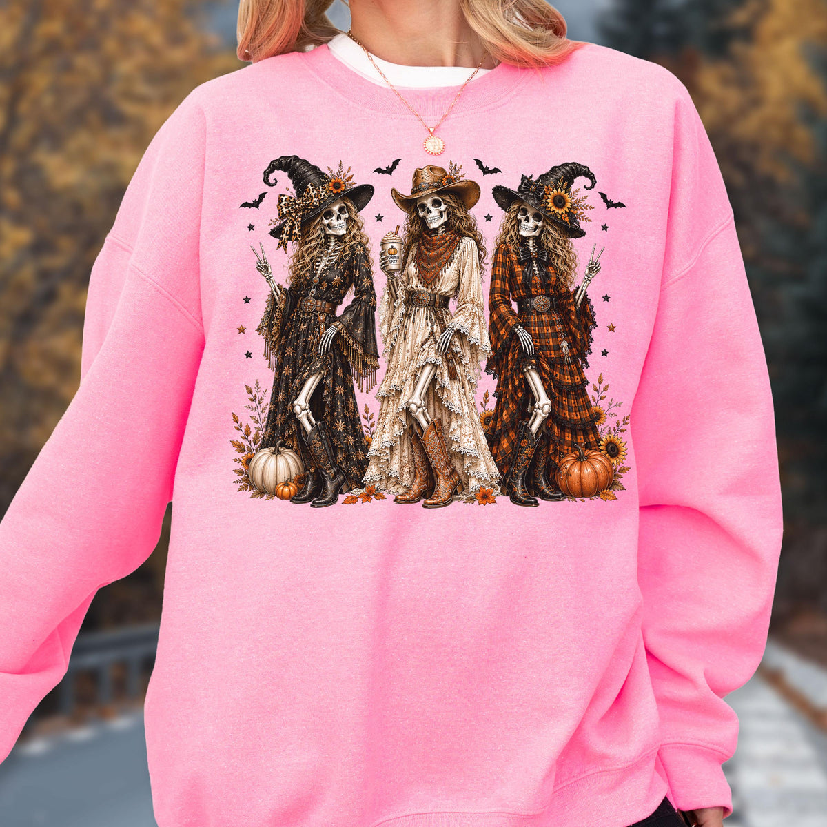 May include: A bright pink crewneck sweatshirt featuring a graphic print of three skeleton figures dressed in long dresses, hats, and boots. The skeletons are surrounded by pumpkins, sunflowers, and small bats, with a white necklace visible above.