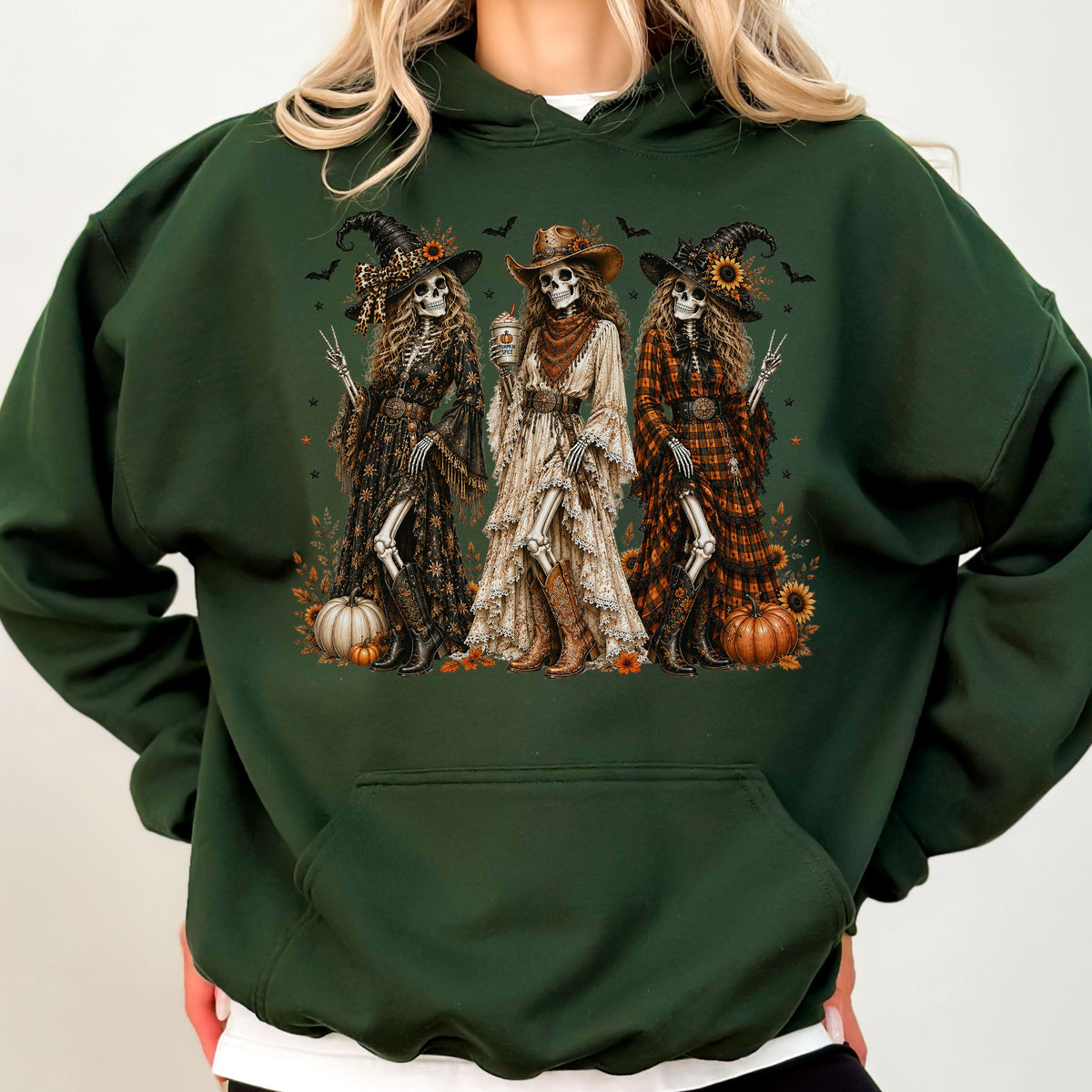 May include: A dark forest green hooded sweatshirt featuring a graphic print of three skeletons dressed in western-style clothing, including hats, long dresses, and boots. The skeletons are surrounded by small pumpkins, sunflowers, and flying bats.
