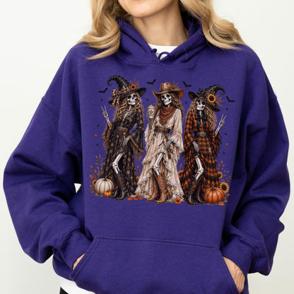 May include: A deep purple hooded sweatshirt featuring a graphic print of three skeletons dressed in western-style clothing, hats, and boots. The skeletons are surrounded by sunflowers, pumpkins, and small bats against a dark, festive background.