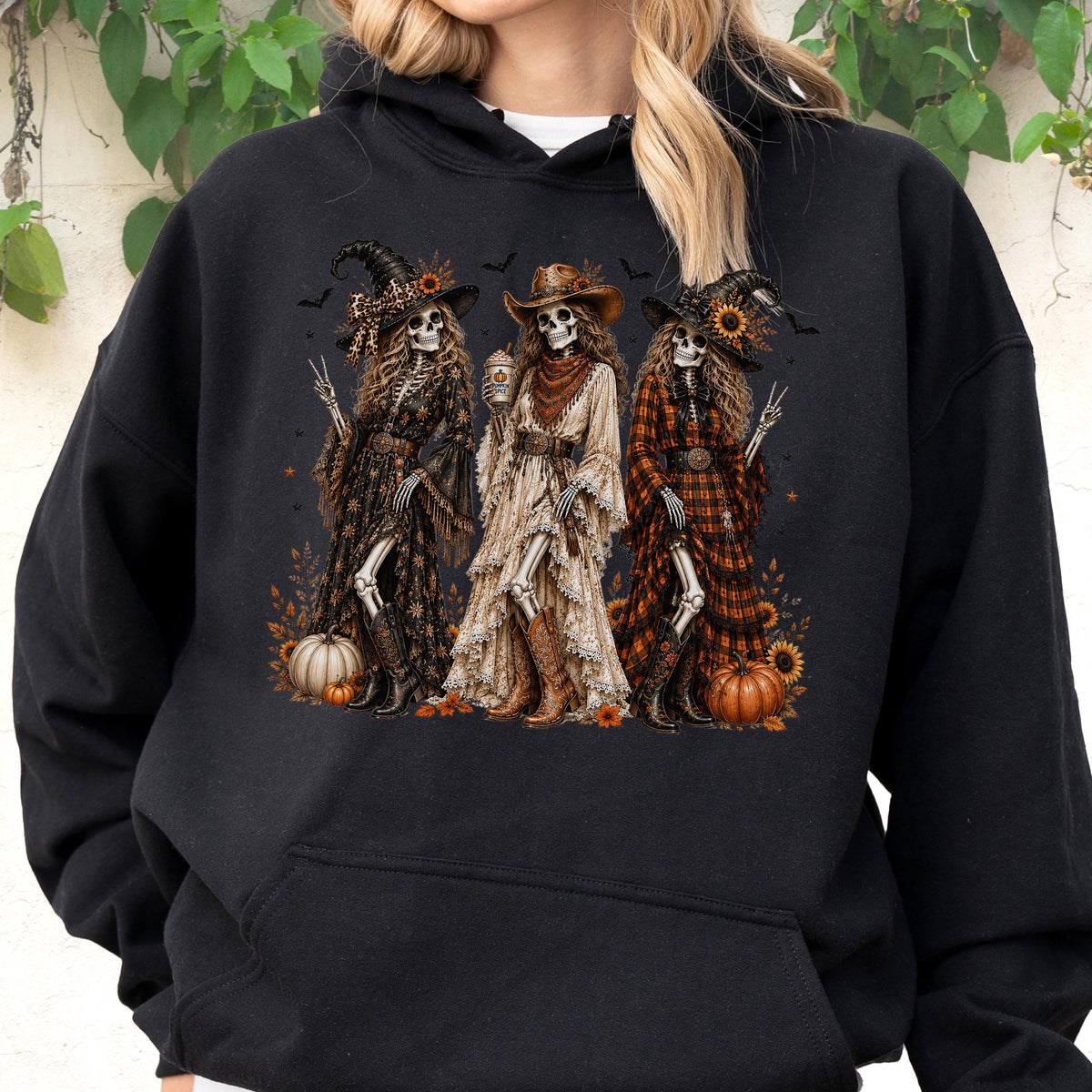 May include: A black hooded sweatshirt featuring a graphic print of three skeletons dressed in western-style clothing, including hats, long dresses, and boots. The skeletons are surrounded by pumpkins, sunflowers, and small bats on a dark background.