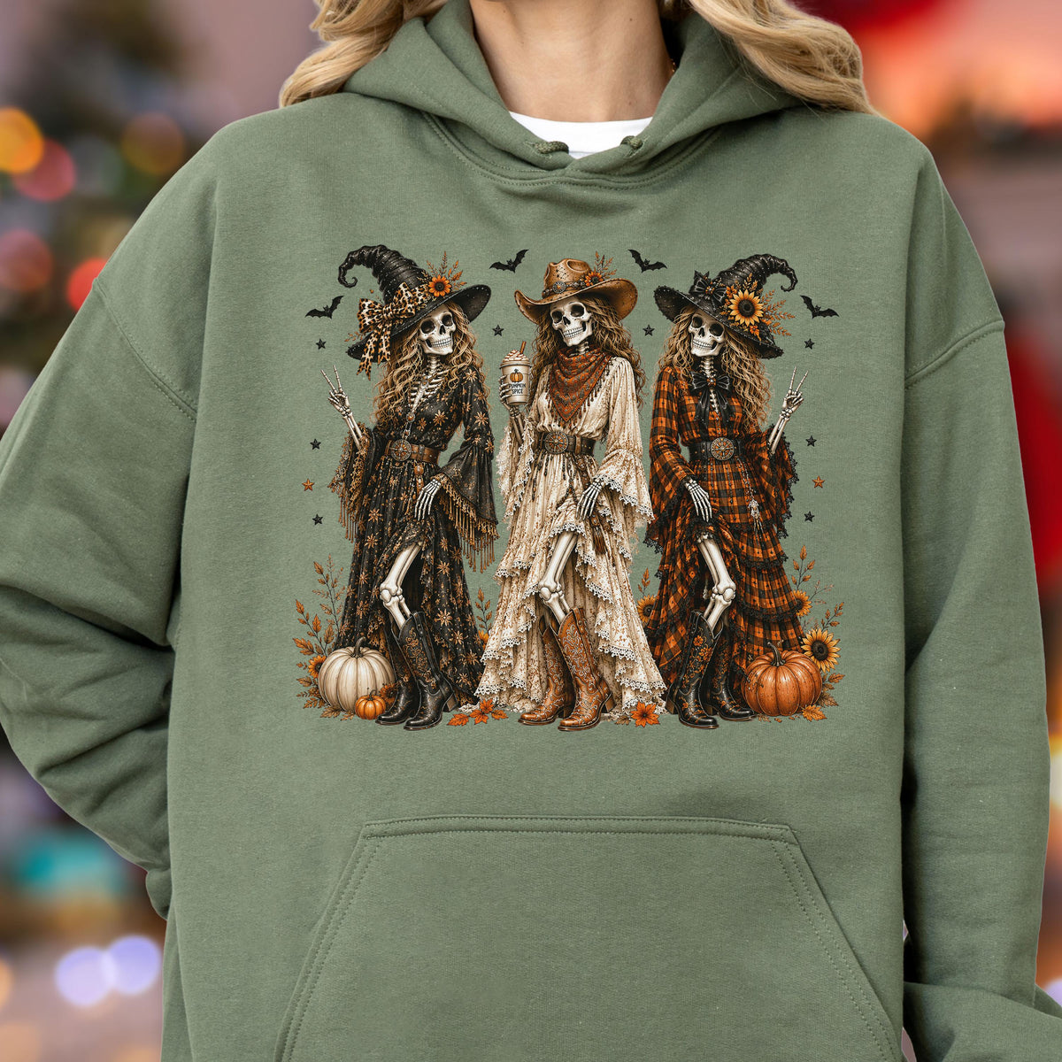 May include: An olive green hooded sweatshirt featuring a graphic print of three skeletons dressed as cowgirl witches. The skeletons wear hats, long dresses, and boots, surrounded by pumpkins, sunflowers, and small bats on a dark background.