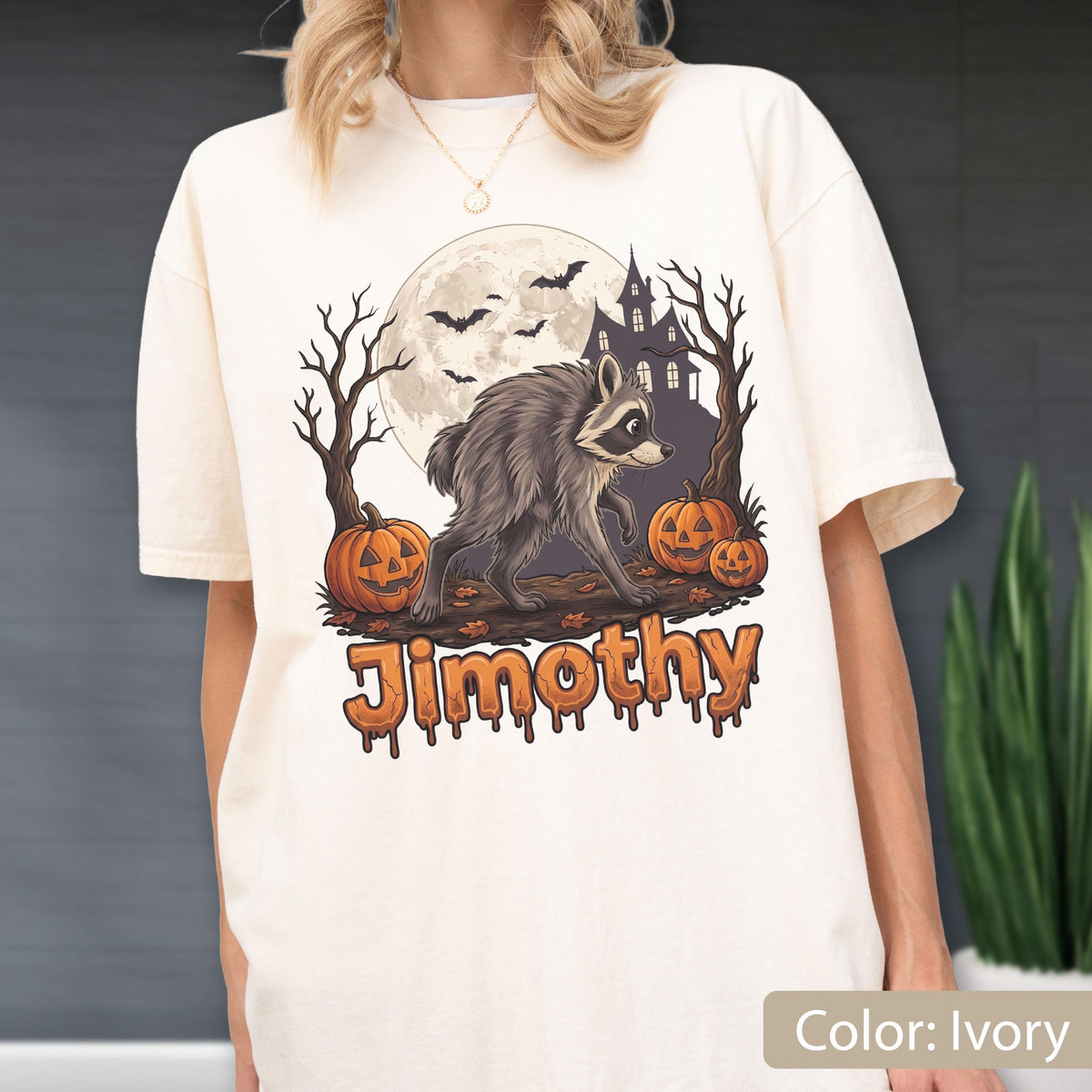 May include: An ivory t-shirt featuring a graphic of a raccoon walking in front of a full moon, a haunted house, bare trees, and two jack-o'-lanterns. The name Jimothy is printed below the illustration in an orange, dripping, bubbly font.