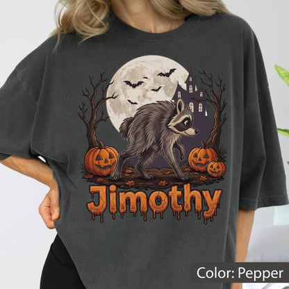 May include: A pepper-colored t-shirt featuring a graphic of a raccoon walking in front of a full moon, a haunted house, bare trees, and three jack-o'-lanterns. The text "Jimothy" is printed in a dripping, orange, pumpkin-textured font below.