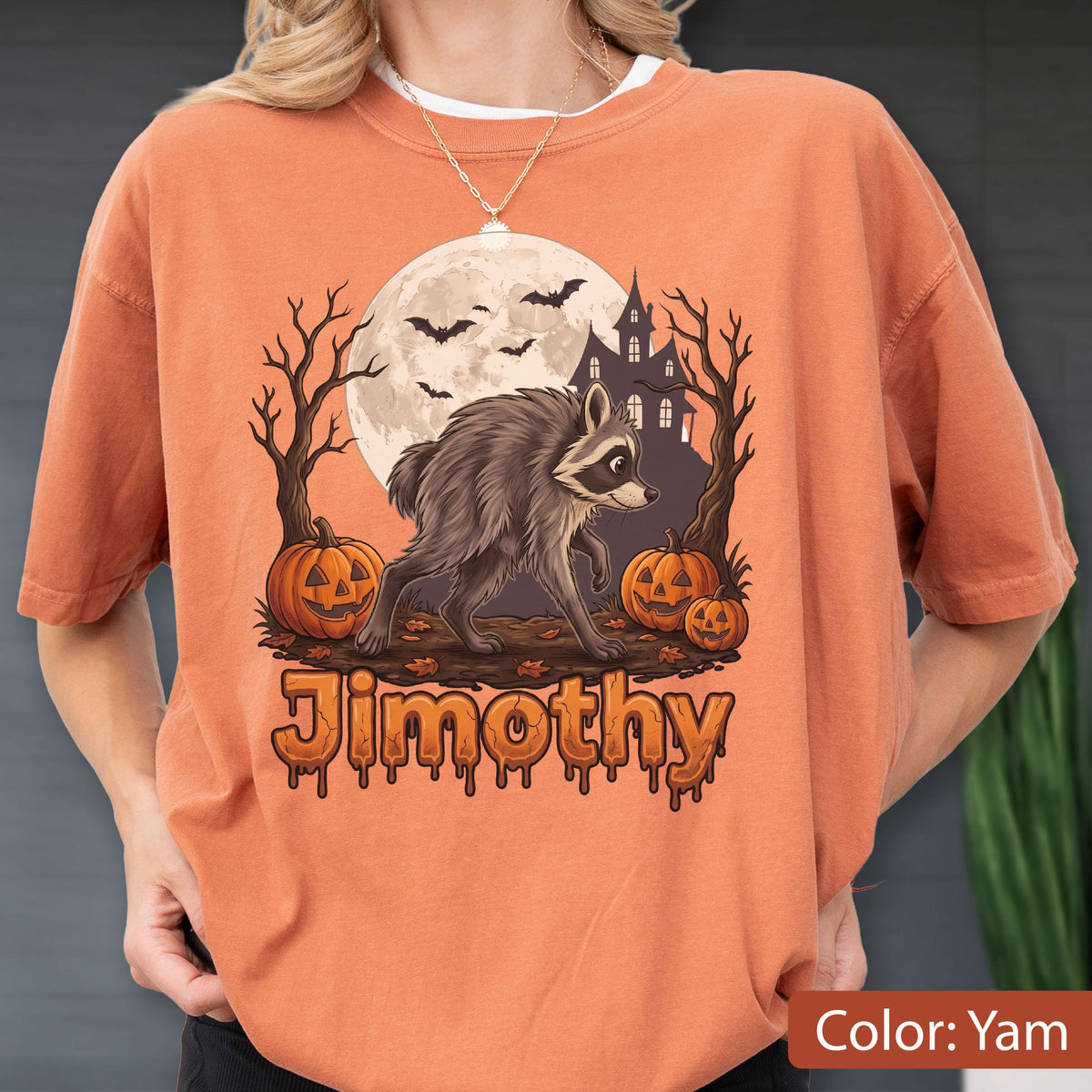 May include: An orange t-shirt featuring a graphic of a raccoon standing in front of a full moon, a haunted house, bare trees, and two jack-o'-lanterns. The text "Jimothy" is printed in a dripping, orange font at the bottom of the graphic.