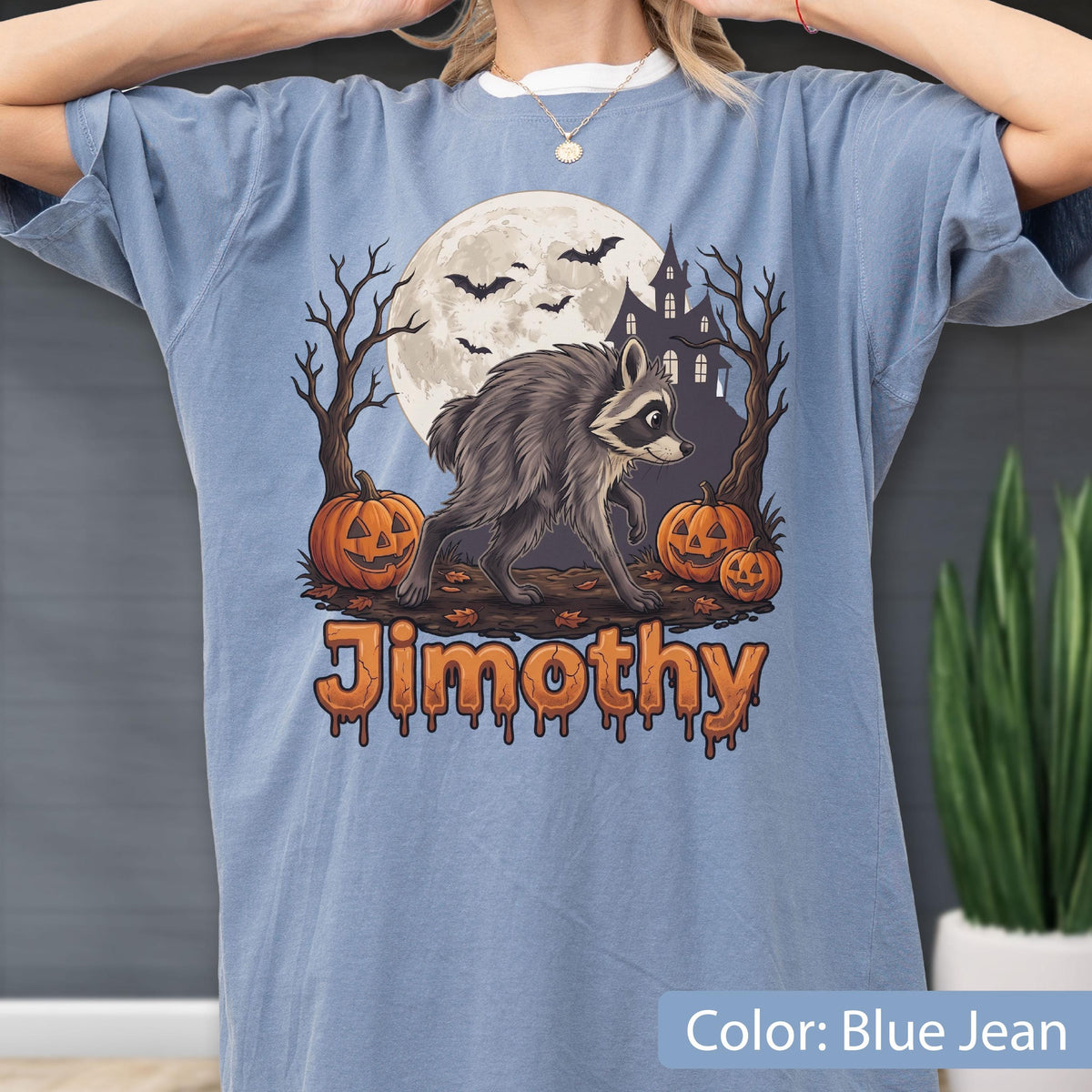 May include: A blue jean colored t-shirt featuring a graphic of a raccoon standing between two jack-o'-lanterns, bare trees, a full moon, bats, and a haunted house. The name Jimothy is printed in orange, dripping-style lettering below the raccoon.