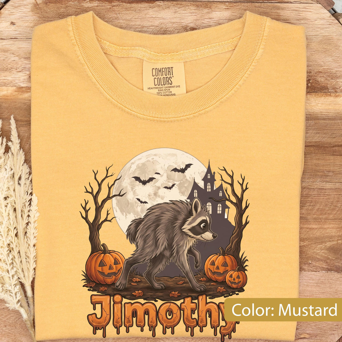 May include: A mustard yellow t-shirt featuring a graphic of a raccoon standing between two pumpkins, a haunted house, and a full moon with bats. The text "Jimothy" is printed at the bottom in a dripping orange font. The shirt has a Comfort Colors label.