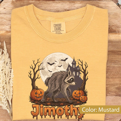 May include: A mustard yellow t-shirt featuring a graphic of a raccoon standing between two pumpkins, a haunted house, and a full moon with bats. The text "Jimothy" is printed at the bottom in a dripping orange font. The shirt has a Comfort Colors label.