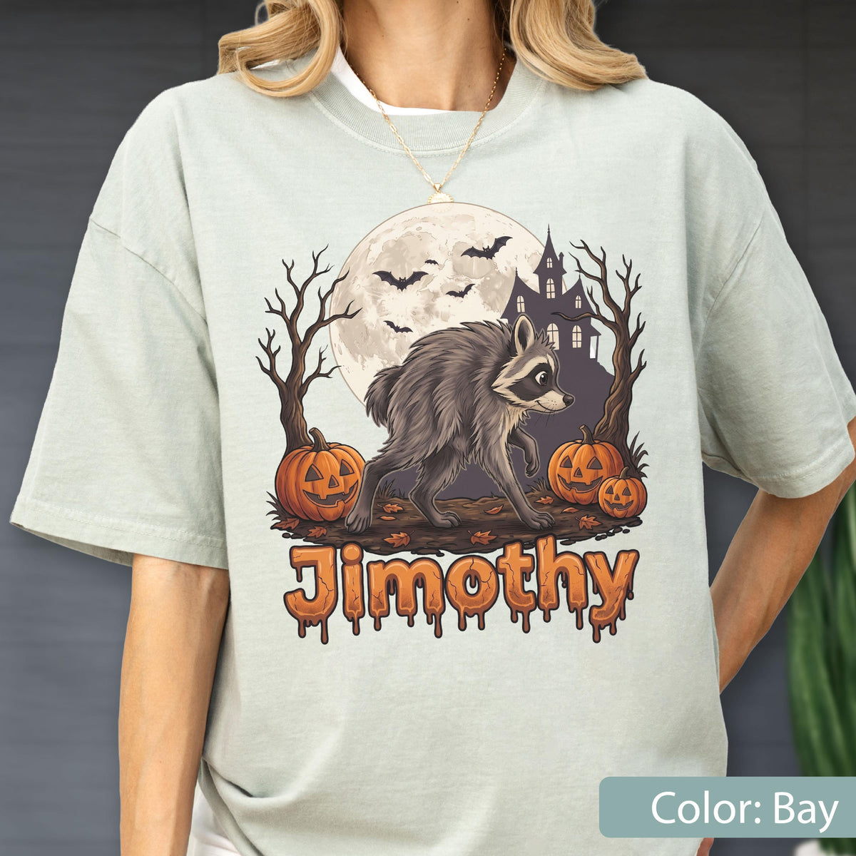 May include: A light sage green t-shirt featuring a graphic of a raccoon walking past two jack-o'-lanterns, bare trees, a large full moon, and a haunted house silhouette. The name Jimothy is printed below in orange, dripping, bubbly lettering.