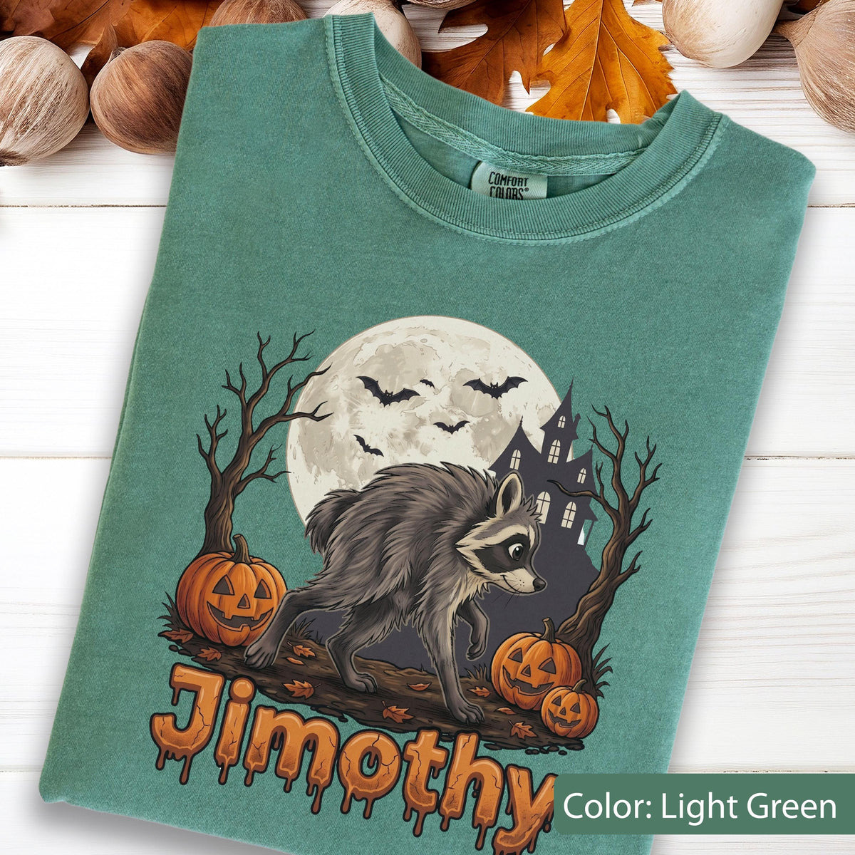 May include: A light green t-shirt featuring a graphic of a raccoon walking past two jack-o'-lanterns, a full moon, bats, and a haunted house. The text "Jimothy" is printed in an orange, dripping font at the bottom of the graphic design.
