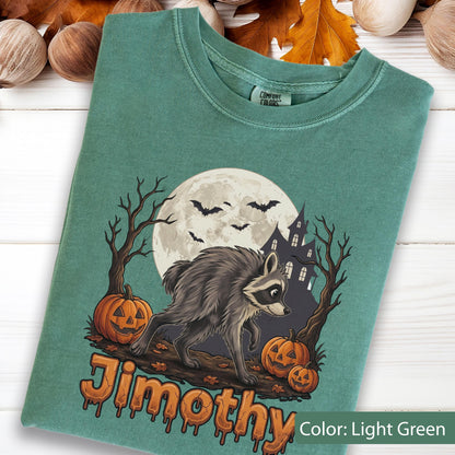 May include: A light green t-shirt featuring a graphic of a raccoon walking past two jack-o'-lanterns, a full moon, bats, and a haunted house. The text "Jimothy" is printed in an orange, dripping font at the bottom of the graphic design.