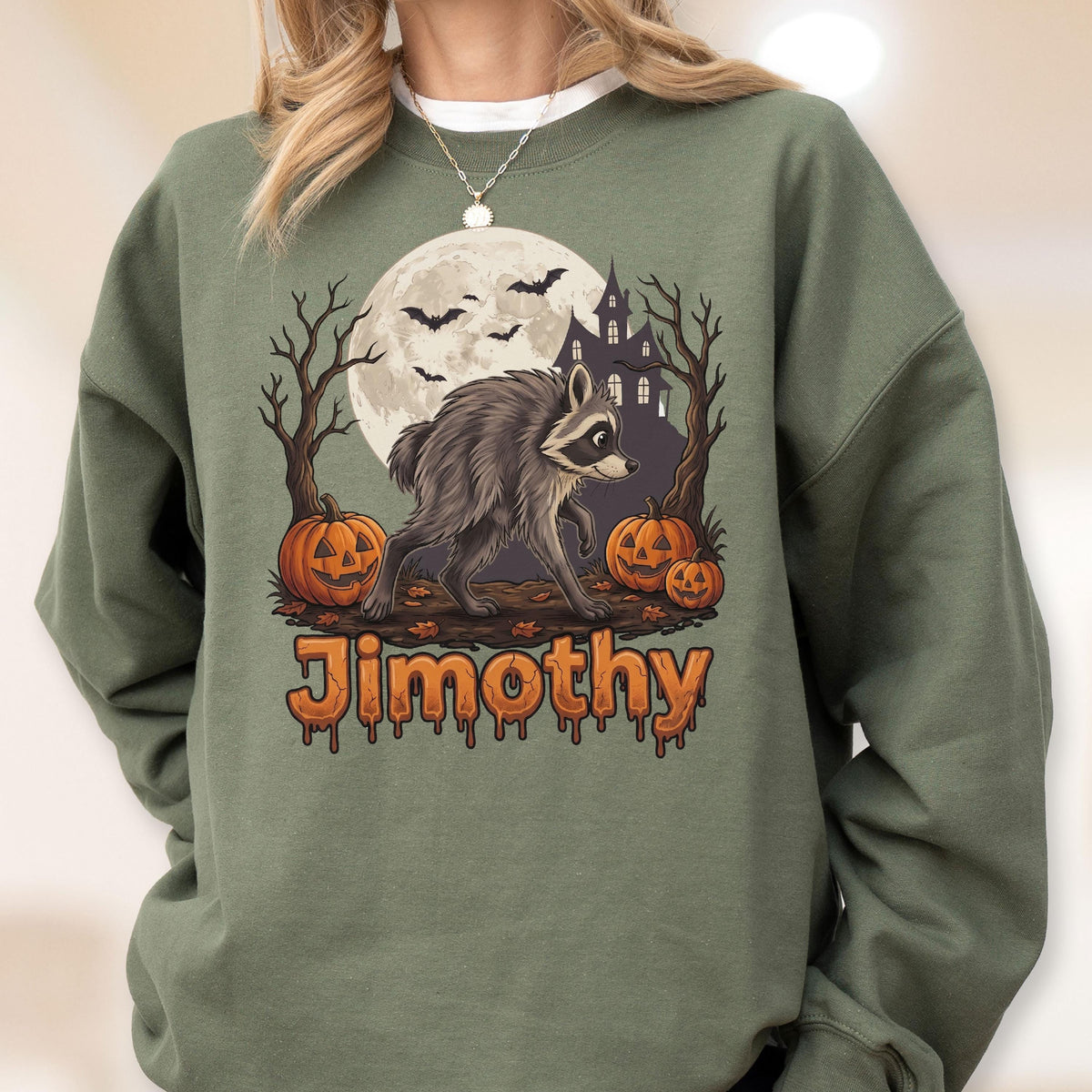 May include: An olive green crewneck sweatshirt featuring a graphic of a raccoon standing in front of a full moon, a haunted house, bare trees, and three jack-o'-lanterns. The name Jimothy is printed in orange, dripping-style lettering below.