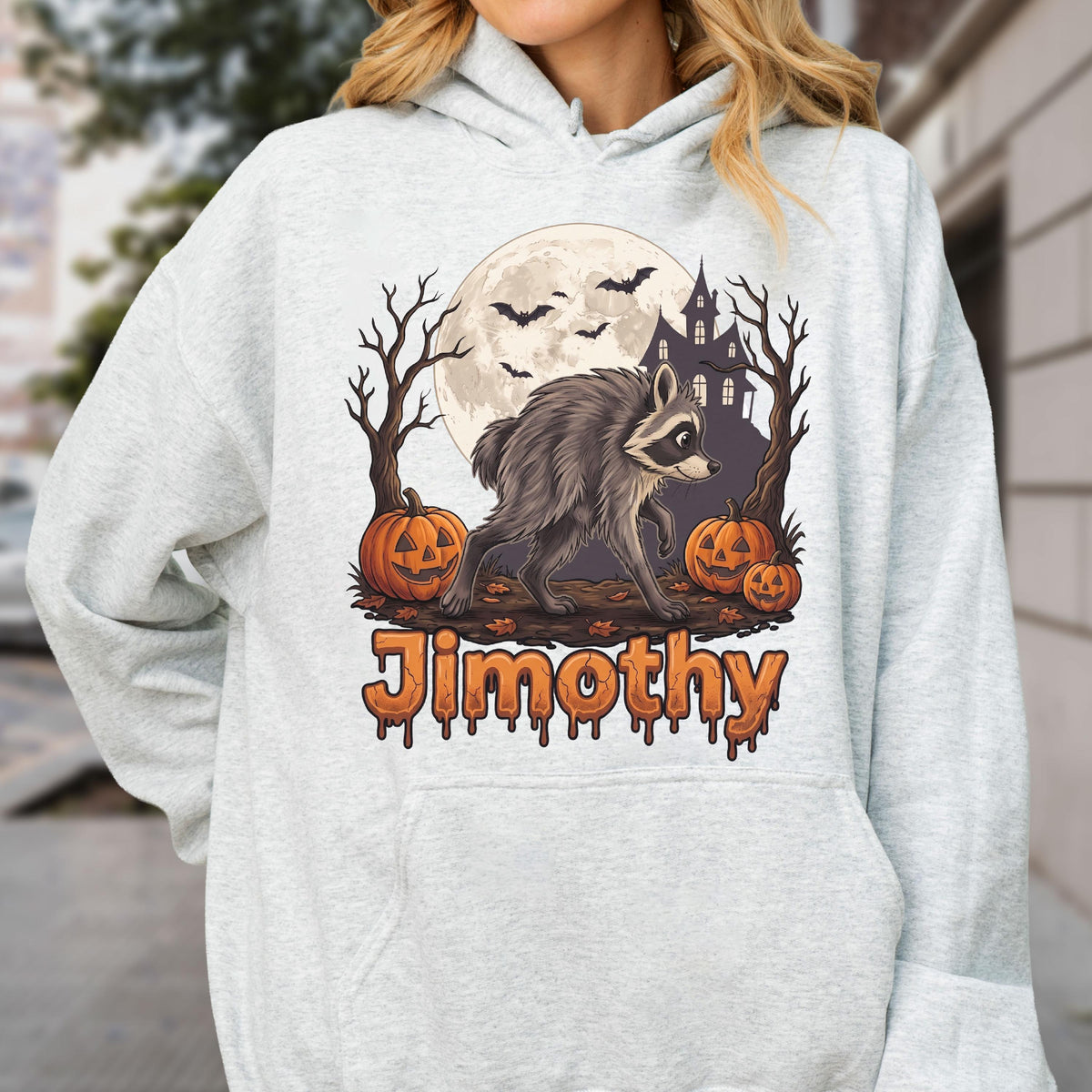 May include: A light gray hooded sweatshirt featuring a graphic of a raccoon walking in front of a full moon, a haunted house, bare trees, and three jack-o'-lanterns. The name Jimothy is printed below the graphic in orange, dripping-style letters.