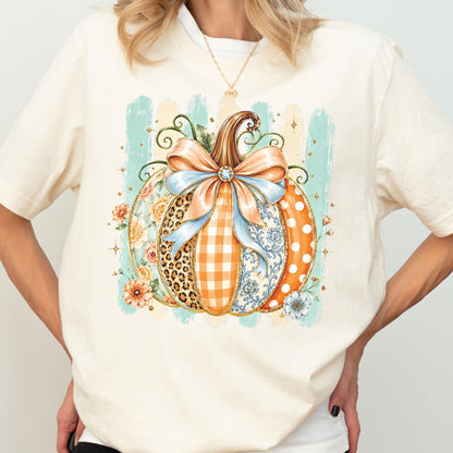 May include: A cream-colored t-shirt featuring a large graphic of a pumpkin decorated with a light blue bow. The pumpkin segments display various patterns including leopard print, orange gingham, floral motifs, and white polka dots on orange.