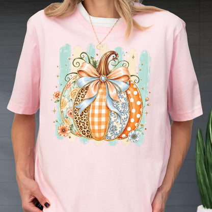 May include: A light pink t-shirt featuring a central graphic of a pumpkin decorated with a large light blue bow. The pumpkin segments display various patterns including leopard print, orange gingham, floral motifs, and white polka dots on orange.