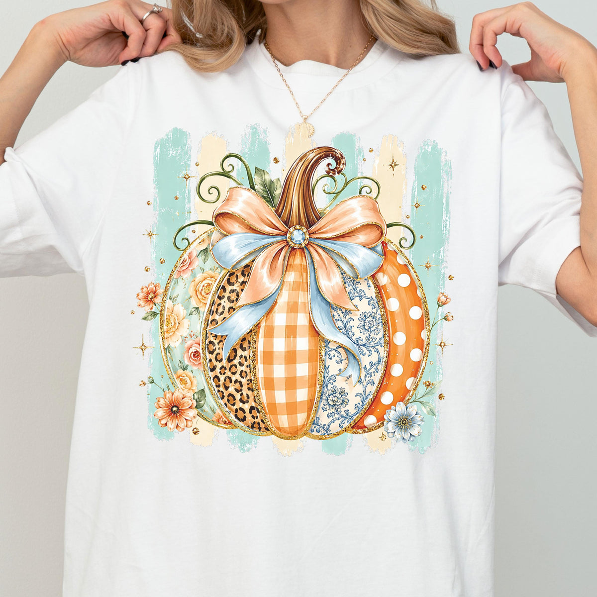 May include: A white t-shirt featuring a large central graphic of a pumpkin decorated with various patterns including leopard print, orange gingham, blue floral, and white polka dots. A large light blue bow sits atop the pumpkin.