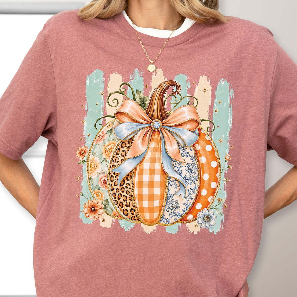 May include: A dusty rose t-shirt featuring a central graphic of a pumpkin adorned with a large light blue bow. The pumpkin segments display varied patterns including leopard print, orange gingham, floral motifs, and white polka dots on orange.