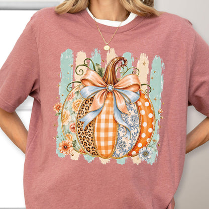 May include: A dusty rose t-shirt featuring a central graphic of a pumpkin adorned with a large light blue bow. The pumpkin segments display varied patterns including leopard print, orange gingham, floral motifs, and white polka dots on orange.