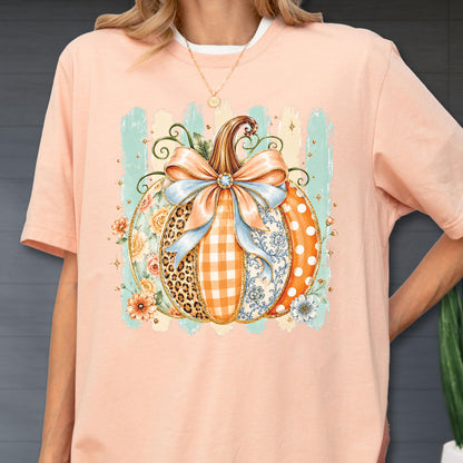 May include: A peach-colored t-shirt featuring a central graphic of a pumpkin decorated with a large light blue bow. The pumpkin segments display various patterns including leopard print, orange gingham, floral motifs, and white polka dots.